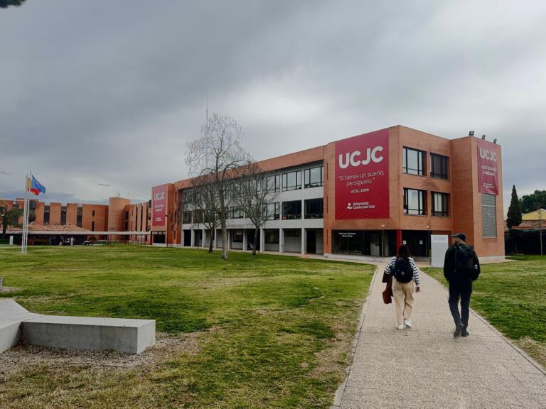 Red brick building of Camilo José Cela University with UCJC banners, seen across a lawn, with two people walking along a path