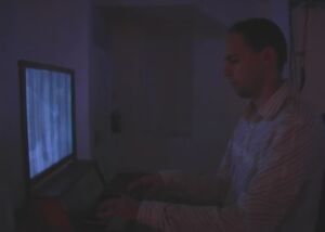Still from a video of Angel of History: a man seated at a keyboard in front of a glowing screen in a dark room