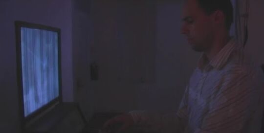 Still from a video of Angel of History: a man seated at a keyboard in front of a glowing screen in a dark room