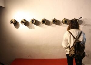 Row of dark shell-casing forms mounted along a white wall, with a visitor looking at them