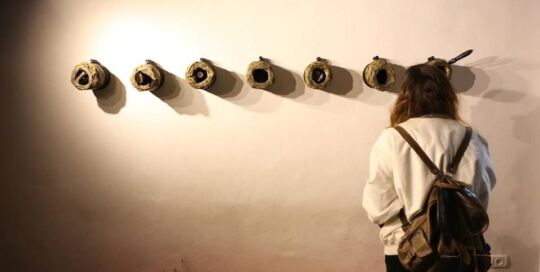 Row of dark shell-casing forms mounted along a white wall, with a visitor looking at them