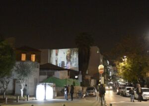 Still from the video Oasis Mosaic, street view: a street at night with a projection glowing on a building wall