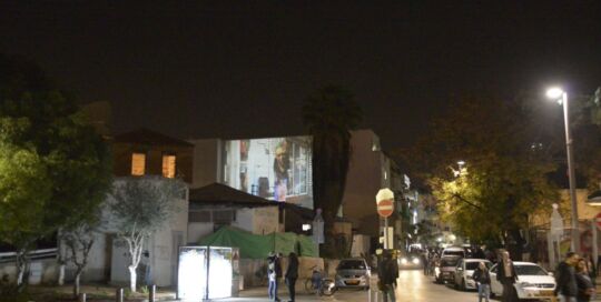 Still from the video Oasis Mosaic, street view: a street at night with a projection glowing on a building wall