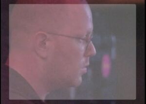 Still from the video Unity Gain: a man with glasses seen on a screen with a pink glow