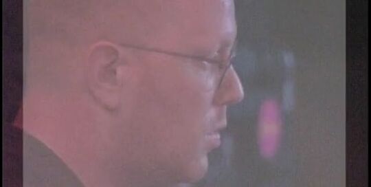 Still from the video Unity Gain: a man with glasses seen on a screen with a pink glow